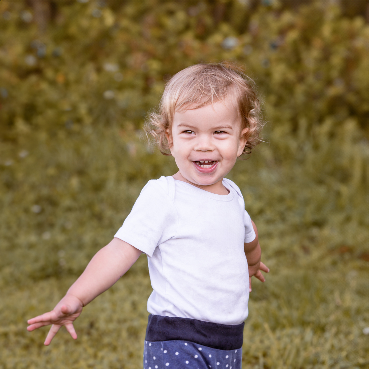 Lachendes Kleinkind mit lockigem Haar läuft im Freien über eine Wiese und streckt spielerisch die Arme aus.